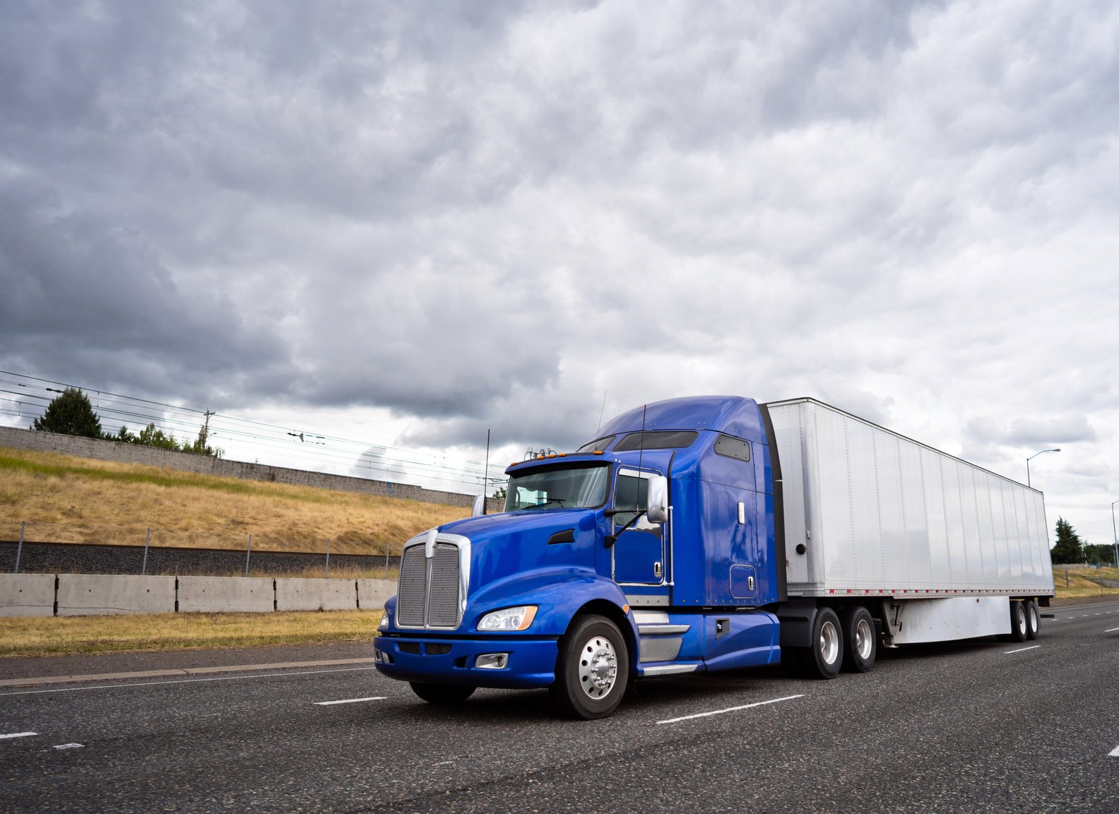 Classic blue big rig semi truck with dry van semi trailer going on wide highway with stormy sky